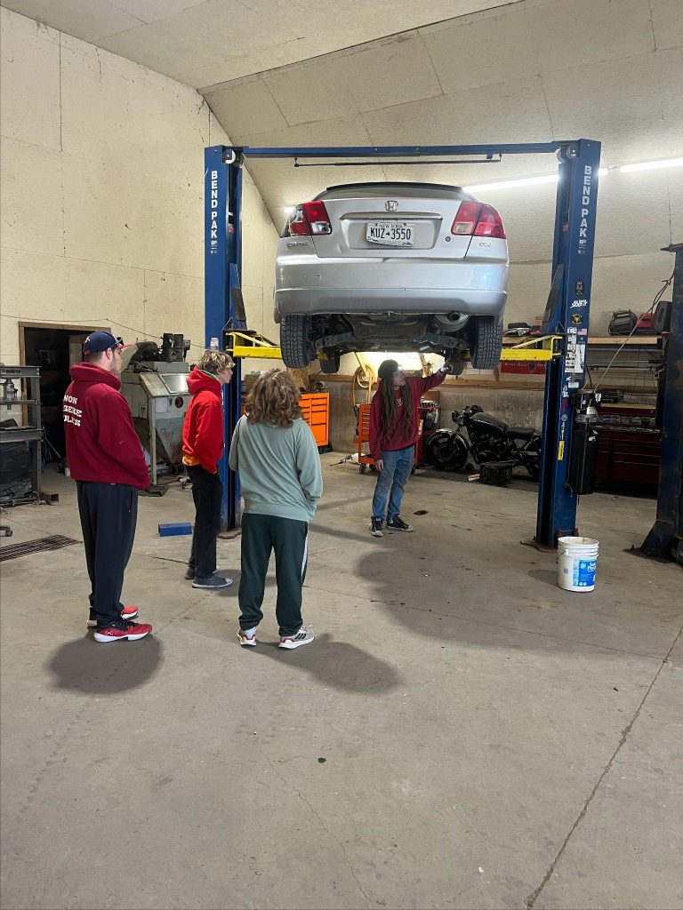 A car is being lifted by a hydraulic lift inside an auto repair shop with people working underneath it and various tools scattered around.