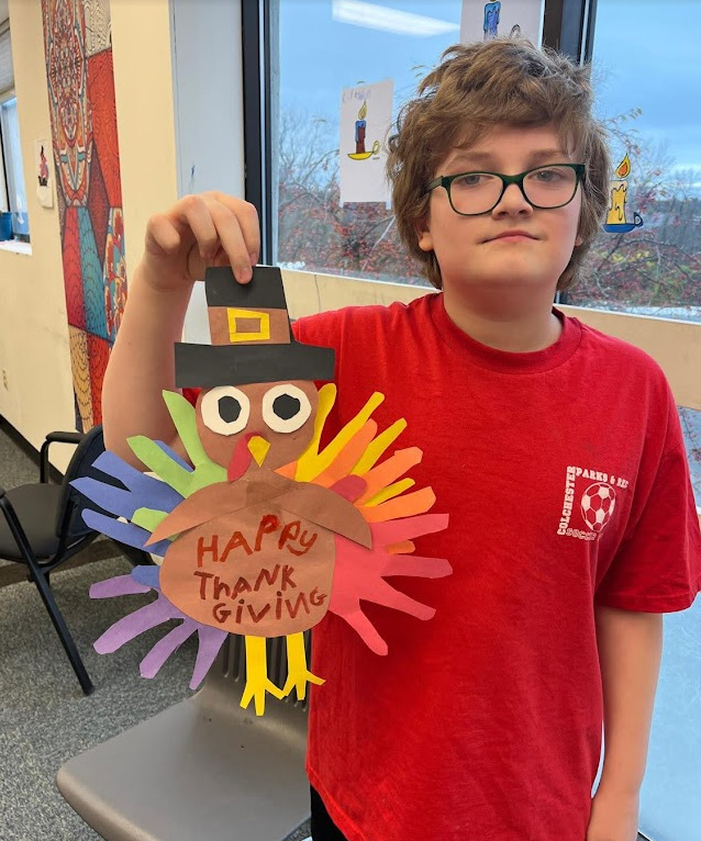 A child is holding a paper cut-out of a turkey with "Happy Thanksgiving" written on it.