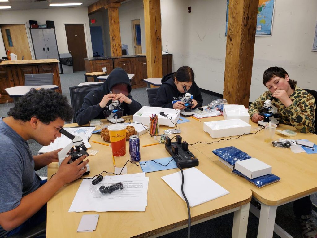 Four individuals are working with microscopes and papers at a table.