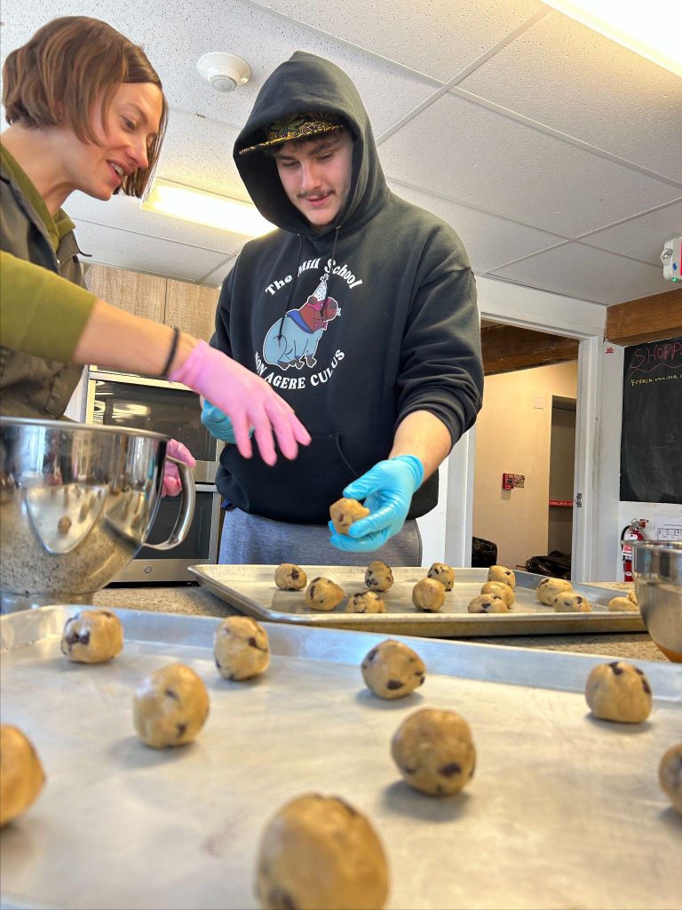 Two individuals are handling dough balls with gloves and a mixing bowl is present.
