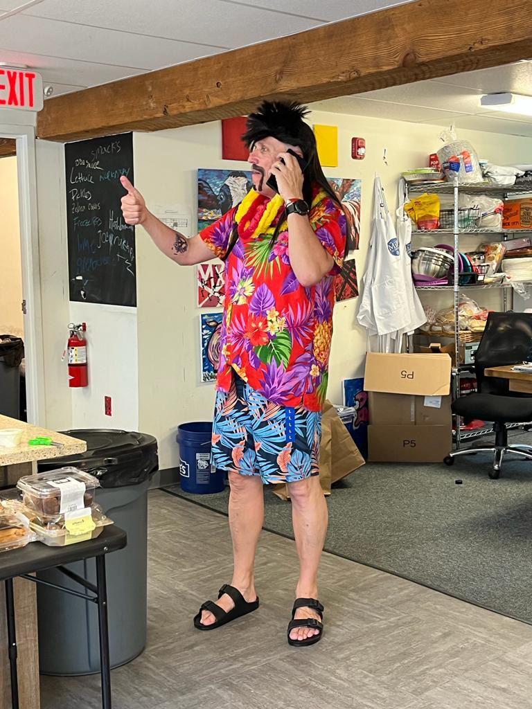 Man with colorful shirt and shorts talking on phone in room with blackboard and shelves.