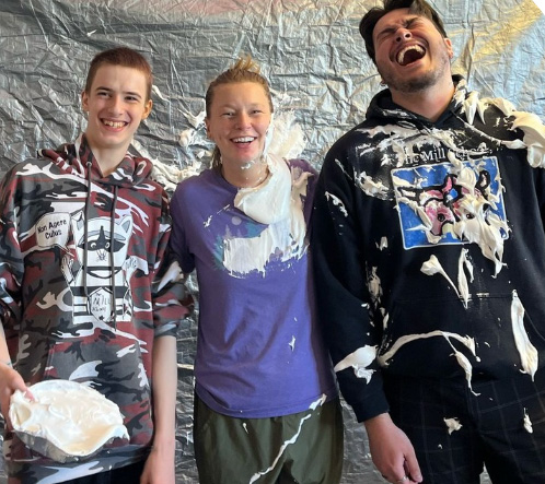 Three young men with white foam covered faces stand against a backdrop of plastic wrap and food remnants.