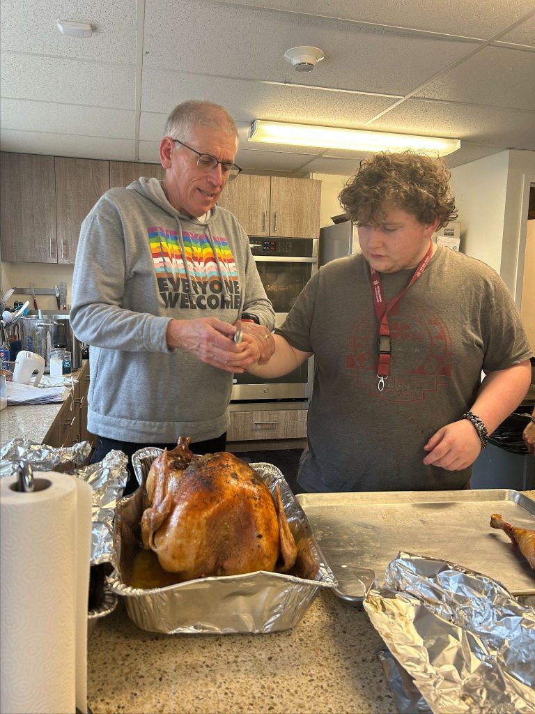 Two men stand behind a table with a roasted turkey and aluminum foil wrapped food items.