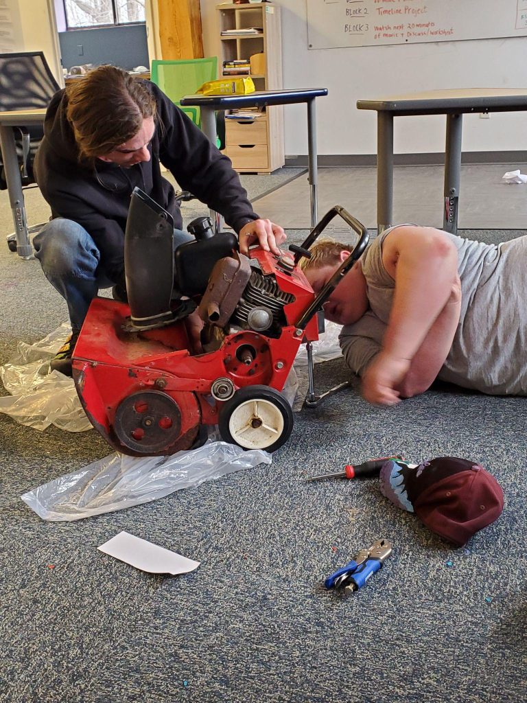 Two men working with a red toy car and tools on the floor.