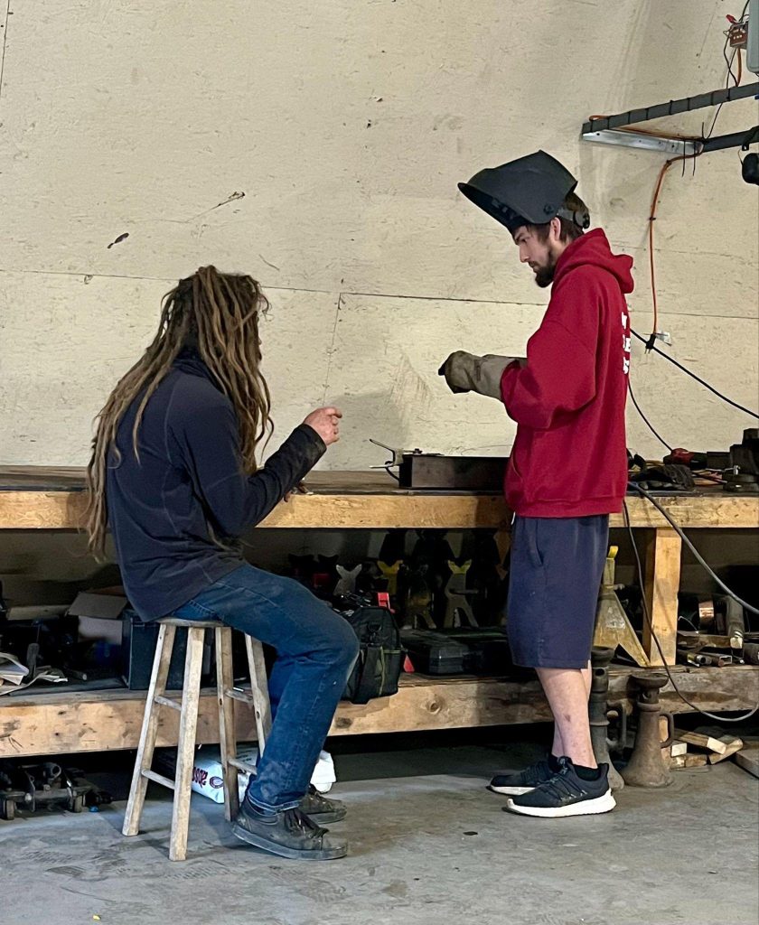 Two men are working at a workbench with tools and equipment behind them.