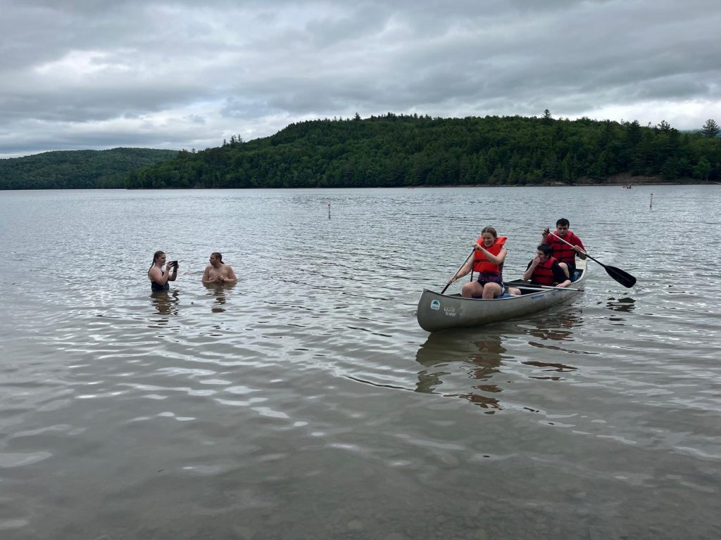 People kayaking near a body of water with mountains and cloudy sky in background.