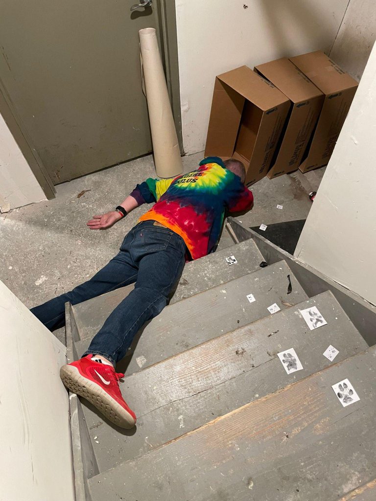 A person is lying face down on a stairway with boxes and playing cards scattered around them.