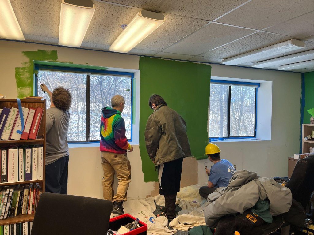 Three people painting a wall with books and clothes scattered around them.