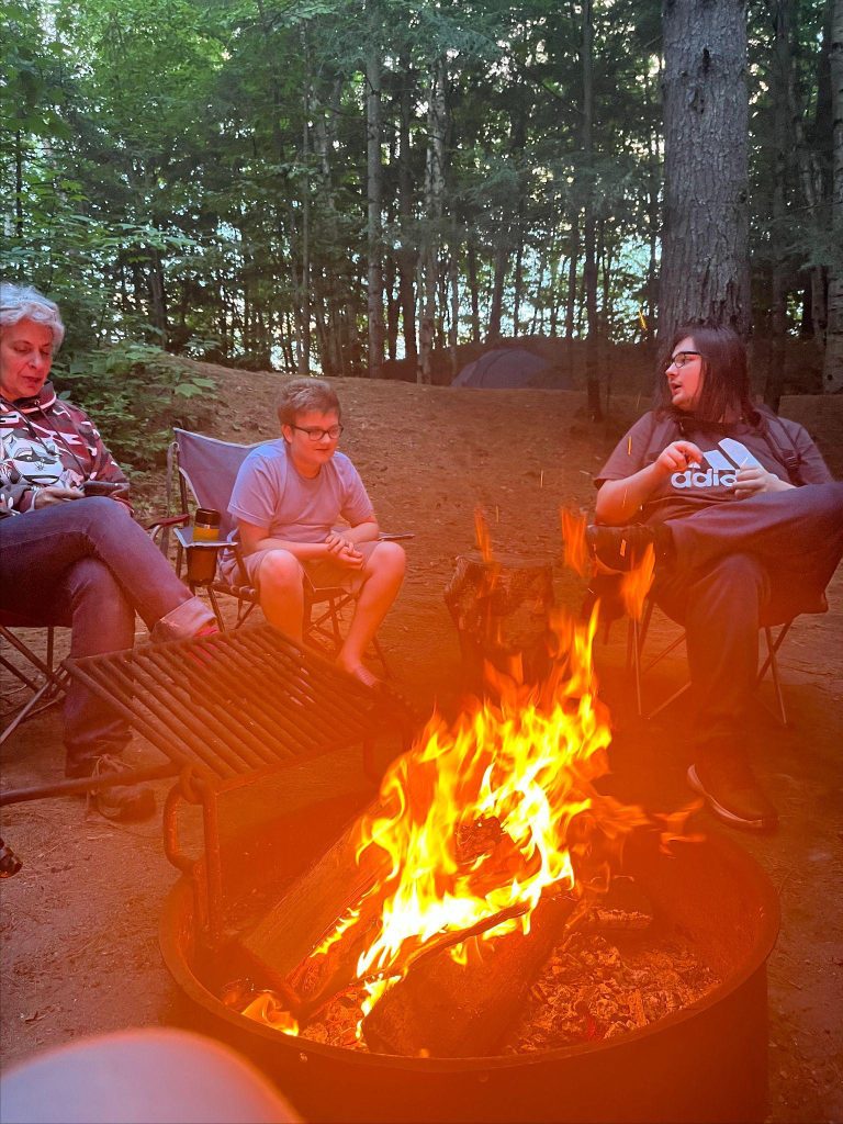 Three people sit around a campfire with flames and smoke rising from it.