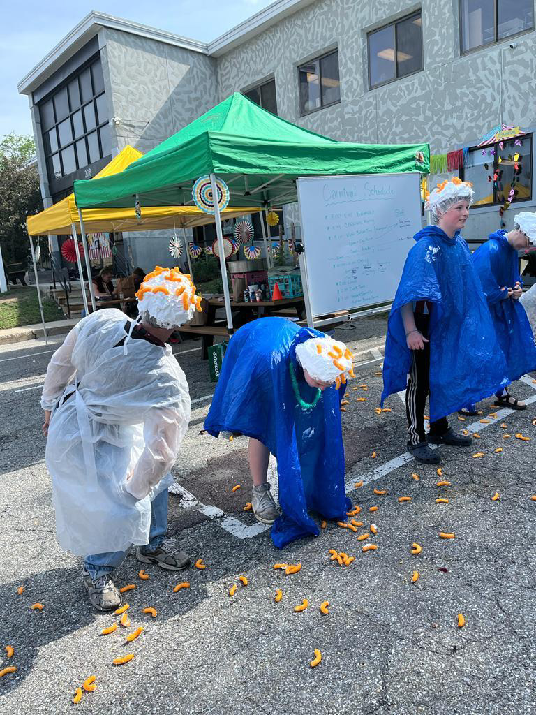 Three people wearing white coats and orange hats are bending over to pick up yellow objects from a street.