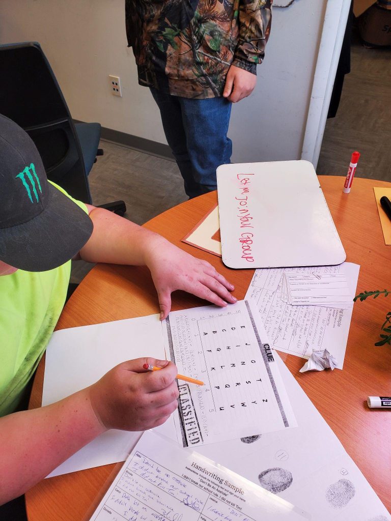 Person is writing with pencil and paper at table while another person stands nearby.