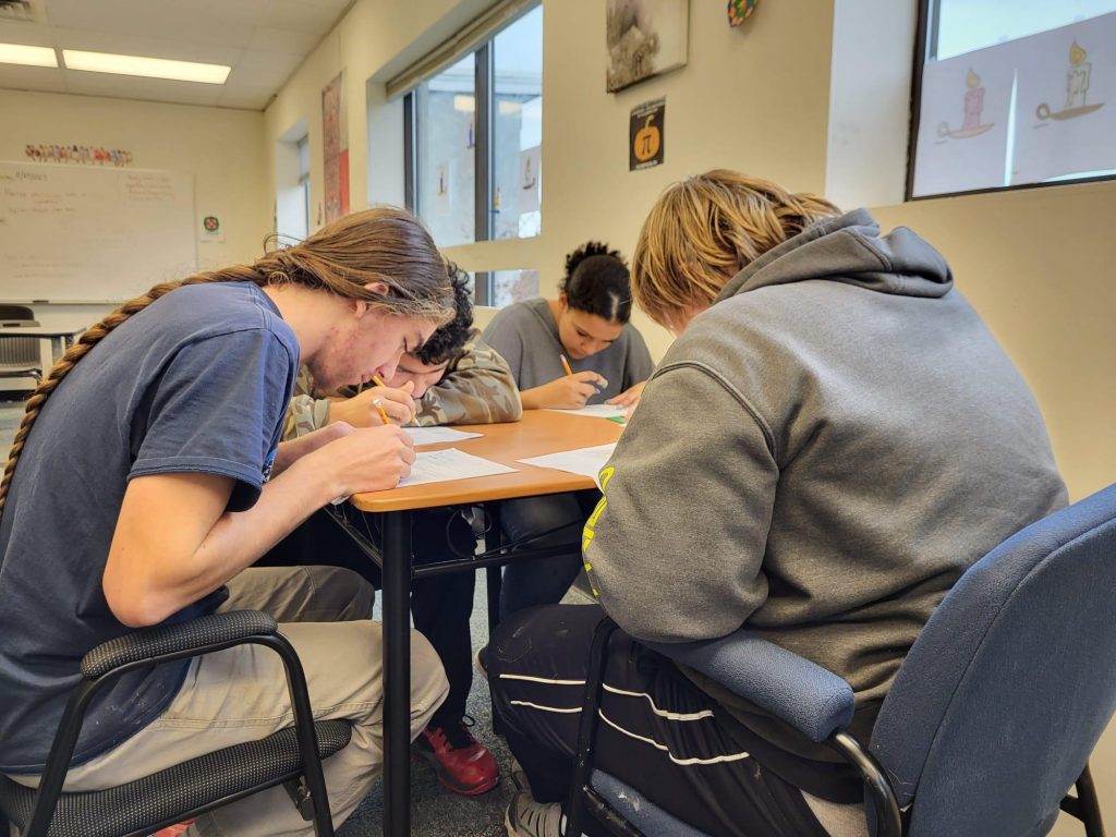 Three students are sitting at a table with papers and writing instruments.